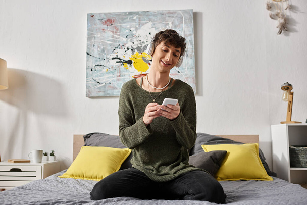 szczęśliwy kręcone transseksualista osoba w bezprzewodowy słuchawki gospodarstwa smartfon i siedzi na łóżku - Zdjęcie, obraz