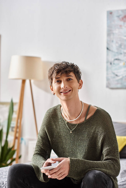 persona transexual feliz en traje casual mirando a la cámara y sentado en apartamento moderno - Foto, imagen