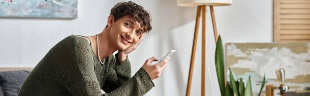 banneri onnellinen kihara transsukupuolinen vaikuttaja älypuhelimella ja istuu makuuhuoneessa, sosiaalinen media - Valokuva, kuva