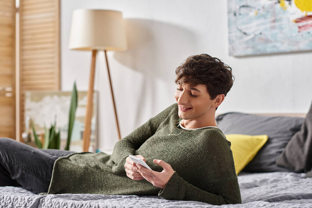 alegre e encaracolado blogueiro transgênero usando smartphone e deitado na cama, influenciador de mídia social - Foto, Imagem