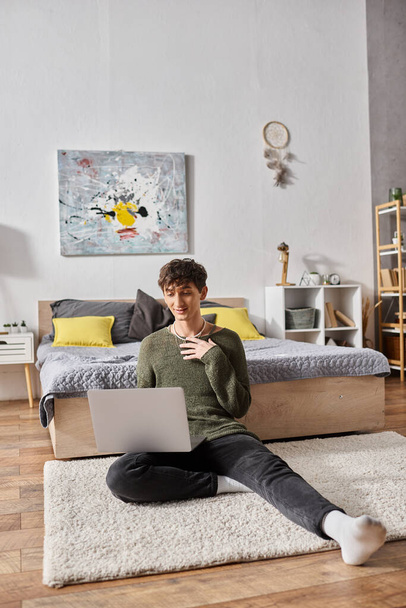 gelukkig en krullend transgender influencer met behulp van laptop en zitten op tapijt in de buurt van bed, blogger - Foto, afbeelding