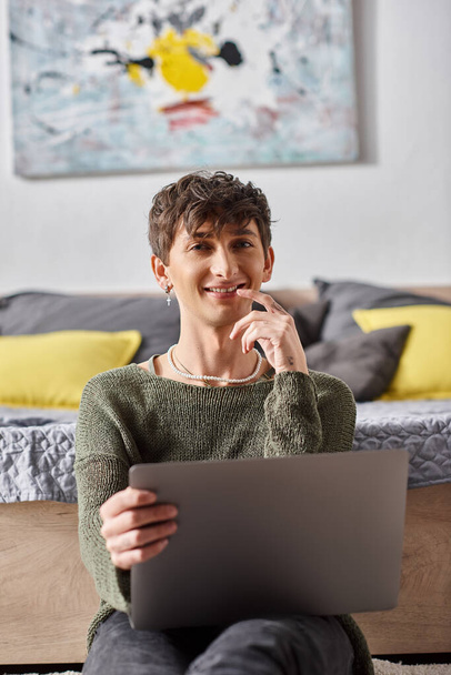 pleased and curly transgender influencer using laptop and sitting next to bed, social media - Photo, Image