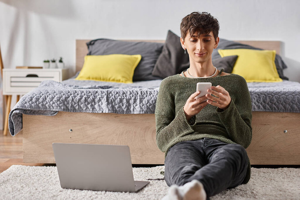 blogger transgénero rizado utilizando el teléfono inteligente junto a la computadora portátil mientras está sentado en la alfombra, las redes sociales - Foto, Imagen