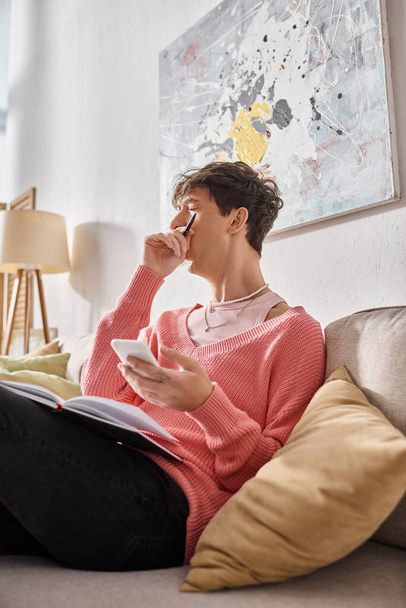 fáradt transznemű blogger rózsaszín pulóverben gazdaság okostelefon és ásítás, miközben ül a kanapén - Fotó, kép