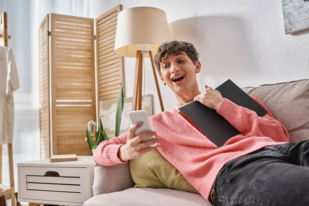 sorprendida persona transgénero en suéter rosa sosteniendo portátil, usando un teléfono inteligente y sentado en el sofá - Foto, imagen