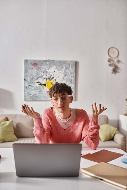 fiatal transznemű szabadúszó rózsaszín pulóverben gesztikulálva, miközben laptopot néz az asztalon, videohívás - Fotó, kép
