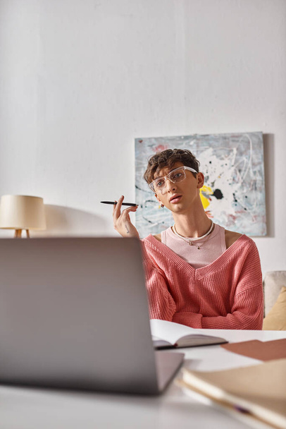 joven transgénero freelancer en suéter rosa gesto mientras mira el ordenador portátil en el escritorio, videollamada - Foto, Imagen