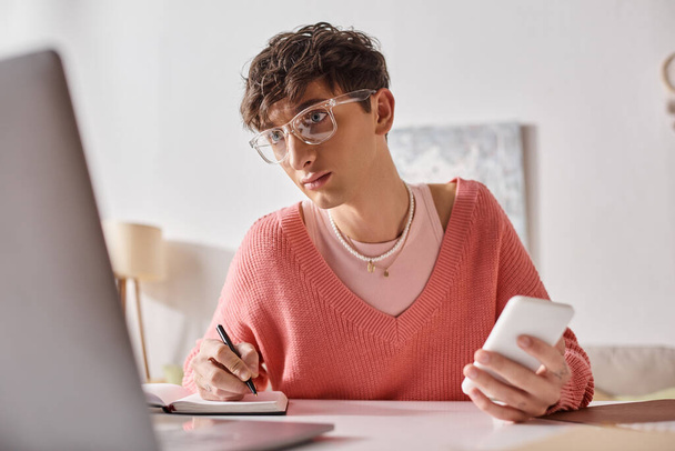 kudrnatý androgynní freelancer v růžovém svetru a brýlích držící smartphone a pomocí notebooku - Fotografie, Obrázek