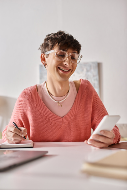 onnellinen androgyyni bloggaaja vaaleanpunaisessa puserossa ja silmälaseissa älypuhelimella ja tekemällä muistiinpanoja - Valokuva, kuva