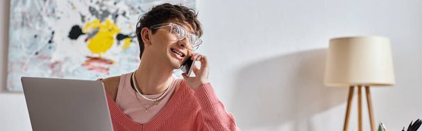 cheerful androgynous freelancer in pink sweater and eyeglasses using laptop and smartphone, banner - Photo, Image