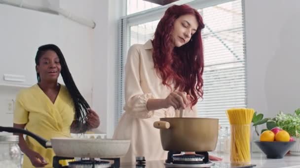 Mediano tiro largo de dos amigas multiculturales hirviendo agua en la cacerola en la estufa mientras cocinan pasta en casa - Metraje, vídeo