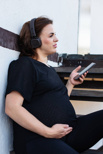Pregnant woman with smartphone listening music in headphones outdoor on white background, spending free time relaxing. Technology, motherhood, mental health, stress relief, mediation and pregnancy. - Photo, Image