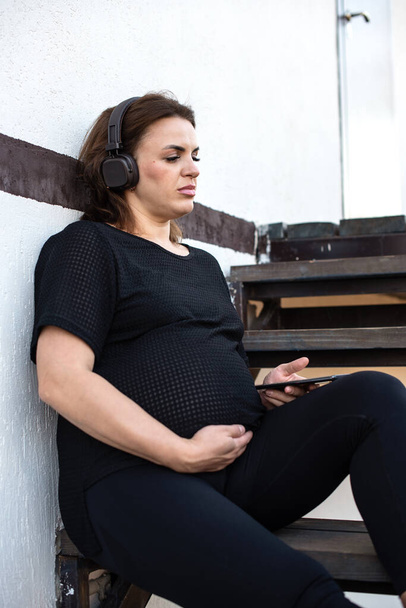 Έγκυος γυναίκα με smartphone ακούγοντας μουσική σε ακουστικά εξωτερική σε λευκό φόντο, ξοδεύοντας ελεύθερο χρόνο χαλαρώνοντας. Τεχνολογία, μητρότητα, ψυχική υγεία, ανακούφιση από το άγχος, διαμεσολάβηση και εγκυμοσύνη. - Φωτογραφία, εικόνα