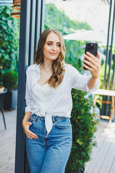 cute young woman in jeans and a white casual shirt is talking on the phone. Video communication via telephone. Remote freelance work. - Photo, Image