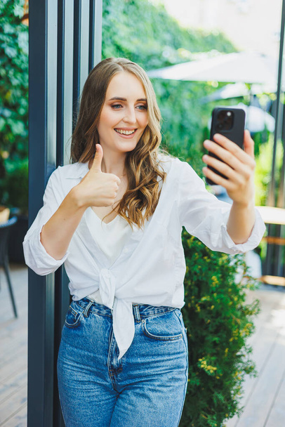 Χαριτωμένη νεαρή γυναίκα με τζιν και ένα λευκό casual πουκάμισο μιλάει στο τηλέφωνο. Τηλεοπτική επικοινωνία μέσω τηλεφώνου. Απομακρυσμένη ανεξάρτητη εργασία. - Φωτογραφία, εικόνα