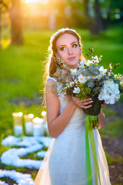 Портрет середньої довжини красивої молодої стрункої сексуальної білої нареченої в модній модній білій сукні, що тримає свіжий квітковий букет, що стоїть на прикрашеному зовнішньому місці в підсвічуванні, дивлячись вгору ліворуч - Фото, зображення