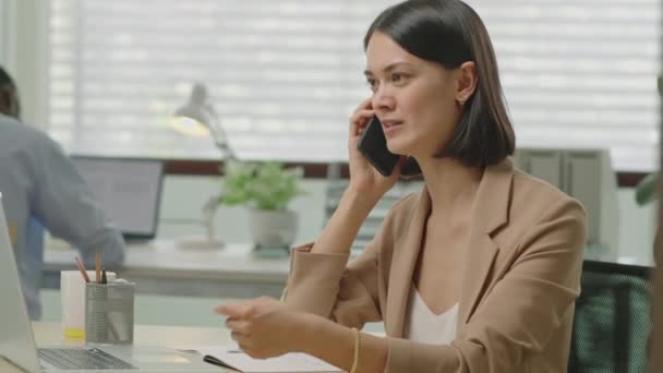 Средний снимок молодой профессиональной предпринимательницы, работающей за столом в офисе и разговаривающей по мобильному телефону - Кадры, видео