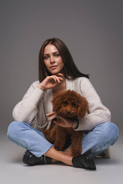 Attractive young brunette in white top and denim shorts, seated on the floor, cradling her brown toy poodle, against a gray studio backdrop - Photo, Image