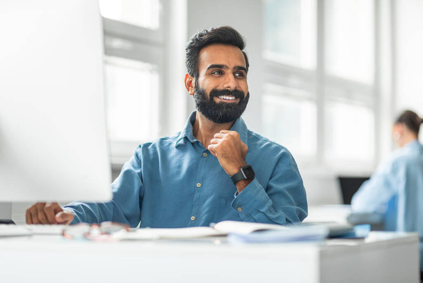 Alegre trabajador de oficina indio trabajando en la computadora y sonriendo mirando a un lado, sentado en el lugar de trabajo en la oficina. Administrador exitoso y concepto de carrera empresarial - Foto, Imagen
