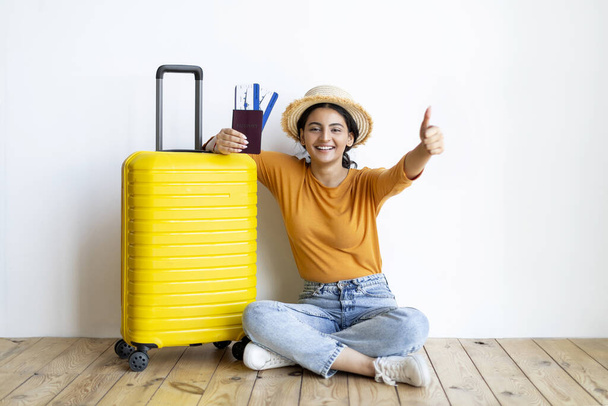 Счастливая молодая индийская женщина позирует с чемоданом и билетами на самолет дома и показывает большой палец вверх, взволнованная восточная женщина в Соломенной шляпе готова к путешествию в отпуск, сидя на полу рядом с белой стеной в помещении - Фото, изображение
