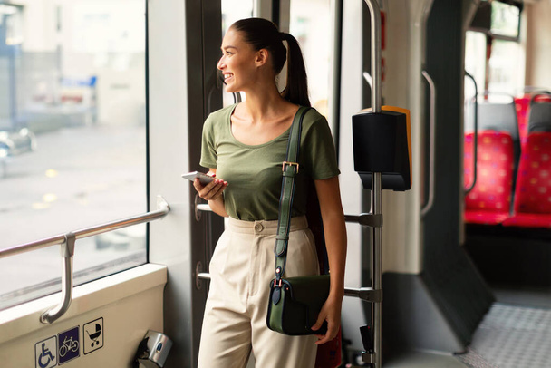 Veselá pasažérka se zapojuje do mobilního telefonu, pohodlně stojí v městské tramvaji. Městský každodenní život s technologií uprostřed tranzitu. Dáma těší sociální média rolování během cesty - Fotografie, Obrázek