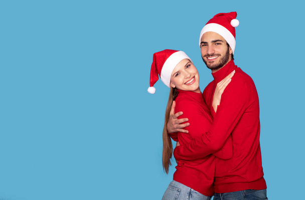 Retrato de abrazar a pareja joven en sombreros de Santa posando sobre fondo azul en el estudio, alegre feliz millennial hombre y mujer abrazando y sonriendo en la cámara, pasando vacaciones de Navidad juntos - Foto, Imagen