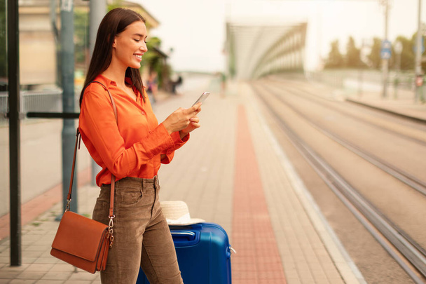 Mladá turistka zasílání zpráv a surfování na webu na mobilním telefonu, čeká na veřejnou dopravu, zatímco stojí s taškou na zavazadla v moderní tramvajové zastávce venku. Volné místo - Fotografie, Obrázek