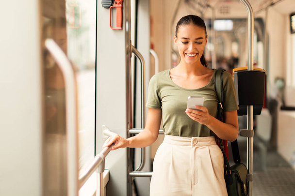 Una joven pasajera sonriente usando su teléfono móvil en un tranvía interior, navegando por la web y reservando billetes de tranvía en línea a través de una aplicación para teléfonos inteligentes. Tecnología digital y transporte moderno - Foto, Imagen