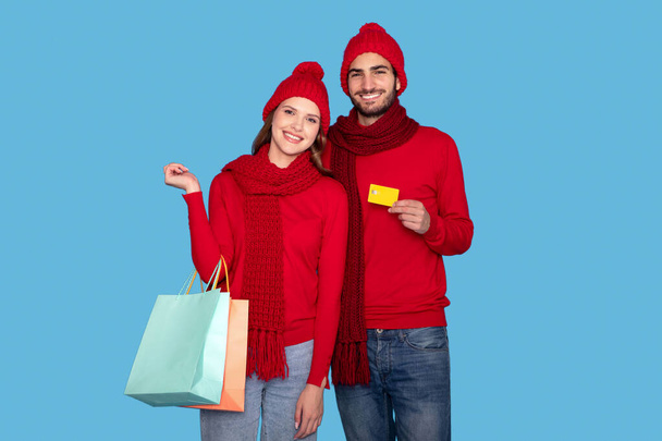 Retrato de casal feliz vestindo chapéus quentes segurando sacos de compras e cartão de crédito, jovem europeu homem e mulher desfrutando de vendas sazonais, de pé isolado sobre fundo azul, espaço de cópia - Foto, Imagem