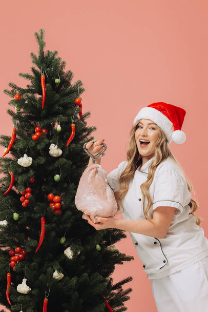Concepto de cocina y vacaciones de Navidad. Una hermosa cocinera rubia en un sombrero de Santa posa sobre el fondo de un árbol de Navidad. - Foto, imagen
