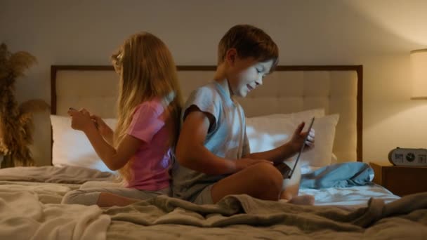 Happy caucasian children boy girl brother sister sibling friends playing online video game together on bed at home evening using laptop and phone young generation internet technology addiction concept - Footage, Video