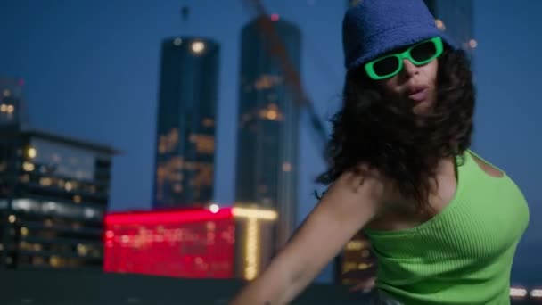 Black young girl with long curly hair moves rhythmically. Sexy fashionable dancer in trendy bucket cup and bright green glasses moves sensually urban downtown. Cool girl dancer dancing in night city - Footage, Video