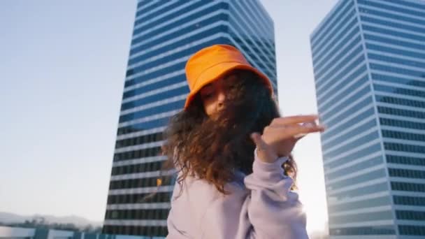 Portrait of young sexy playful multiracial girl dancing in the city, dressed in trendy orange bucket hat with sunset pays light shining between two glass skyscrapers. 30s Woman looking camera close up - Footage, Video