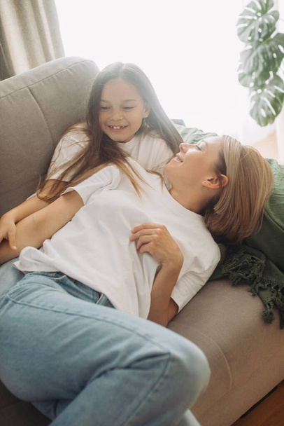 Anyák napja. Egy anya és a lánya játszanak, mosolyognak és ölelkeznek. Anya és lánya otthon ülnek a kanapén egy szabadnapon.. - Fotó, kép