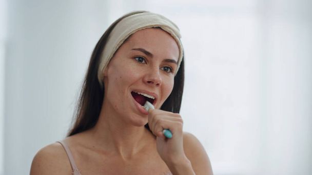 Donna sorridente che fa igiene dentale in bagno da vicino. Bruna donna positiva lavarsi i denti guardandosi allo specchio. Carino ragazza bocca pulita con dentifricio rimozione placca. Concetto quotidiano di routine - Foto, immagini