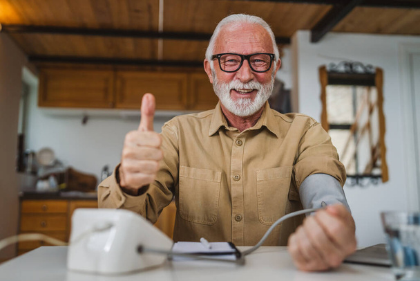 Un uomo felice maschio caucasico anziano con barba sana celebrare utilizzare il dispositivo di pressione sanguigna per controllare e misurare i risultati mentre sedersi a tavola a casa da solo persone reali copiare concetto di assistenza sanitaria spazio - Foto, immagini