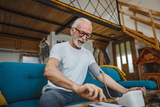 Egy férfi boldog időskorú fehér férfi szakállal egészséges ünneplik használja vérnyomás eszköz ellenőrzésére és mérésére eredményeket, miközben ül az asztalnál otthon egyedül valós emberek másolja tér egészségügyi koncepció - Fotó, kép