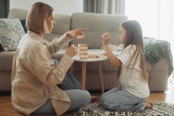 Una madre feliz y su hija pequeña se divierten jugando un juego de mesa. Madre e hija juegan Jenga en la sala de estar. - Foto, imagen