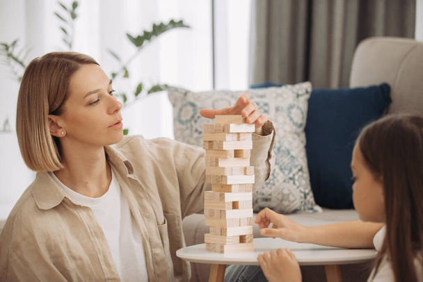 A happy mother and her little daughter have fun playing a board game. Mother and daughter play Jenga in the living room. - Photo, Image