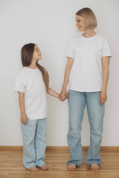 Matka i córeczka ubrane w dżinsy i białe t-shirty pozują na białą ścianę. - Zdjęcie, obraz