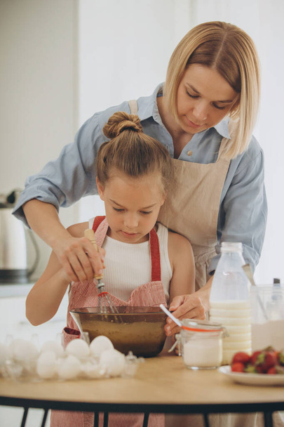 Matka i córka przygotowują ciasto naleśnikowe przy stole w nowoczesnej kuchni. Dziewczyna trzyma trzepaczkę, miesza jajka w misce, przygotowuje ciasto z matką. - Zdjęcie, obraz