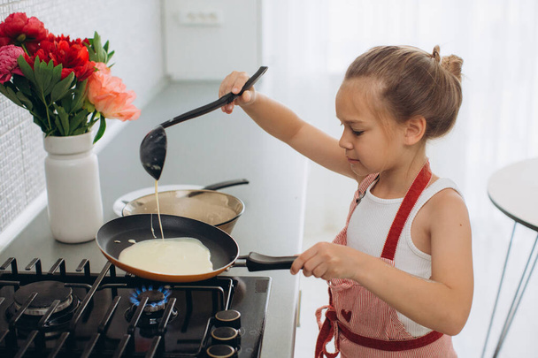 Ein kleines 8-jähriges Mädchen in Schürze brät in der Küche Pfannkuchen. - Foto, Bild