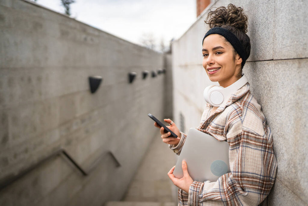 Egy nő fiatal felnőtt nő diák állni vagy sétálni szabadtéri hold laptop számítógép használata okostelefon sms üzenetek online valós személy másolás tér boldog mosoly magabiztos fejhallgató nappal - Fotó, kép