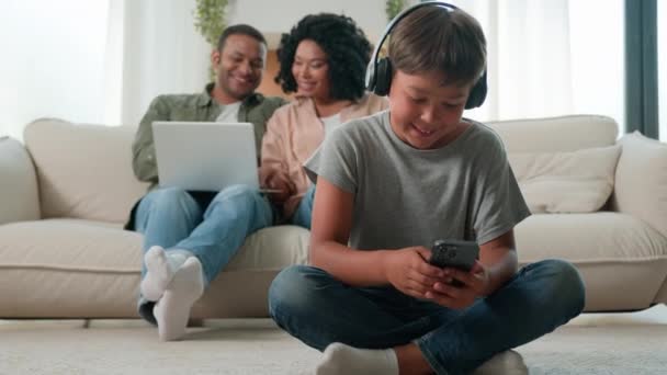 Маленький син хлопчик в навушниках грає у відеоігри на мобільному телефоні щаслива багаторасова мама тато і дошкільнята, використовуючи різні пристрої вдома дорослі батьки купують онлайн з ноутбуком на дивані - Кадри, відео