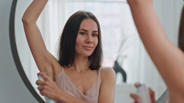 Smiling woman applying antiperspirant at home closeup. Cute lady spraying armpits deodorant enjoying daily procedures. Happy girl looking mirror reflection. Body care daily hygiene routine concept  - Photo, Image