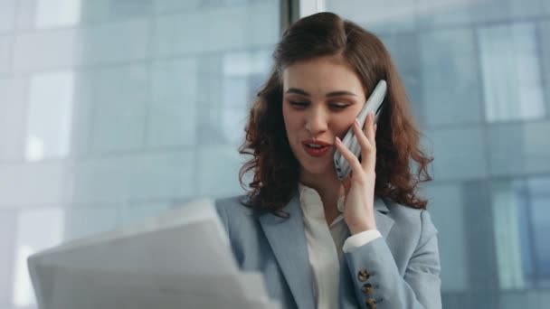 オフィスのマネージャーは,ビジネス文書を閉じている見て笑顔で携帯電話を話します. 現代のオフィスの窓で電話を話す肖像画の魅力的な幸せな女性のコンサルタント. 陽気な女の子は会話を楽しむ. - 映像、動画