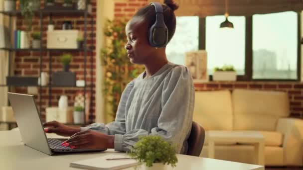 Freelancer werkt aan een bureau met muziek, geniet van online job taken tijdens het luisteren naar liedjes op audio headset. Afro-Amerikaanse vrouw telewerken bij zonsondergang, het schrijven van rapporten over notitieblok. - Video
