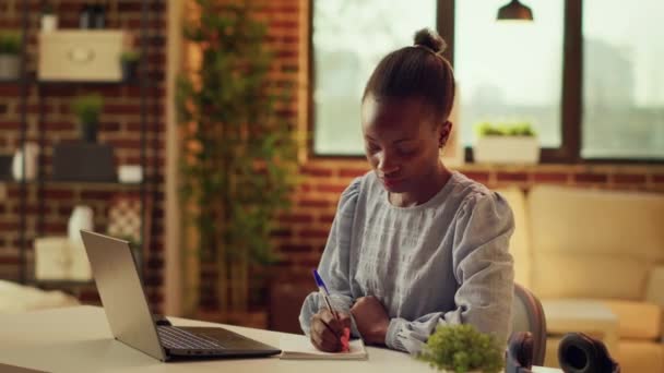 Freelancer taking notes about work, using notebook planner to help with remote job productivity at desk. African american woman writing down ideas as inspiration, home office. - Séquence, vidéo