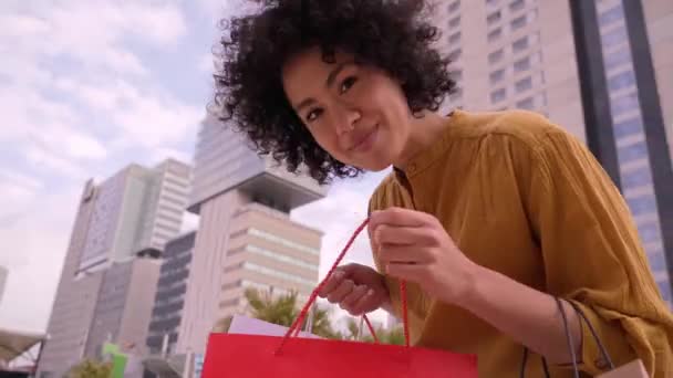 Молодая взрослая латиноамериканка танцует, держа сумки возле торгового центра. Афро-самка представляет счастливый и оживленный внешний вид и демонстрирует покупки. Поколение женщин z, зависимых от сделок - Кадры, видео