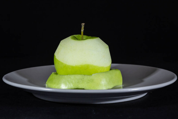 Селективное размытие на зеленом яблоке, наполовину очищенном яблочной кожурой на серой тарелке, изолированной на черном фоне. - Фото, изображение